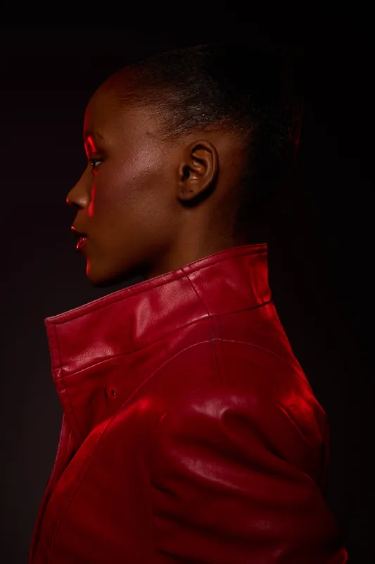 Profile of a dark skinned African woman with natural hair wearing a red leather jacket. Studio lighting highlights her smooth skin and strong features against a black background.