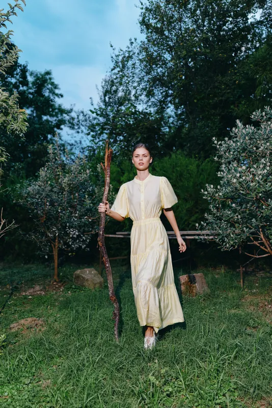 woman in a light dress walks across a grassy garden holding a wooden staff, serene evening light creates a calm atmospheric scene in nature