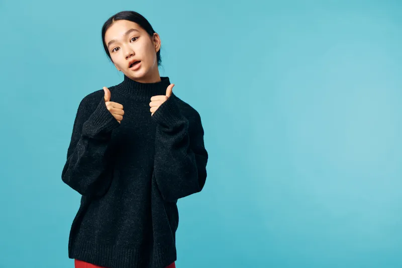 Young woman in black sweater with thumbs up gesture expressing approval and confidence against blue background. Casual style with relaxed pose and direct eye contact.