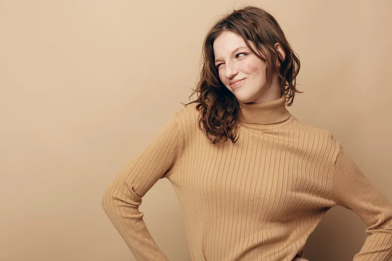 Young woman in beige sweater poses with hands on hips and looks sideways with a slight smile. Neutral background and casual style create a warm, confident mood.
