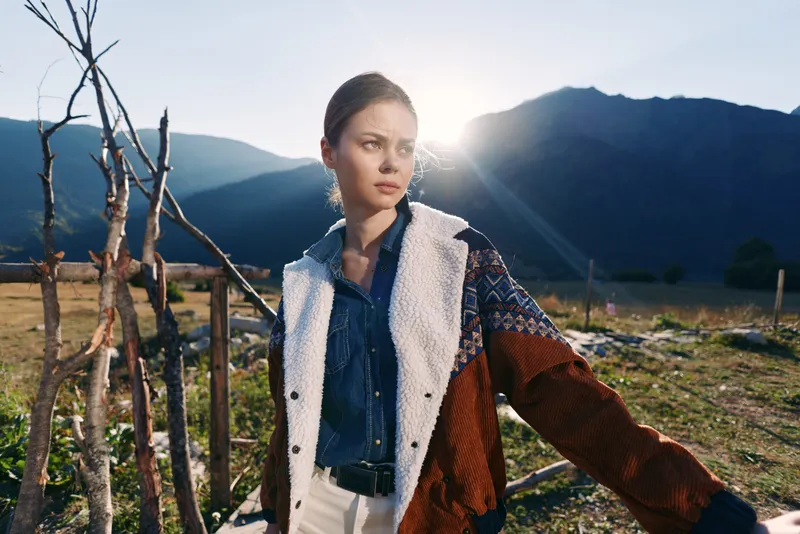Woman portrait in countryside with mountains and sunlight, wearing a shearling jacket and denim shirt by a rustic fence. Outdoor fashion, thoughtful expression and natural scenery.