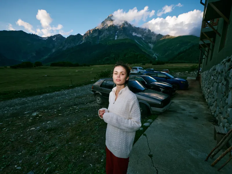 woman in casual sweater stands near parked cars against a rugged valley and distant mountains, capturing a serene outdoor moment with winding roads and expansive landscape