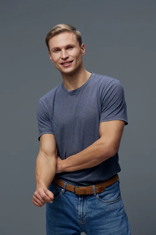 Smiling young man with short blond hair wearing casual blue t-shirt and jeans, standing against plain gray background, cheerful expression, casual style, lifestyle concept, studio portrait.