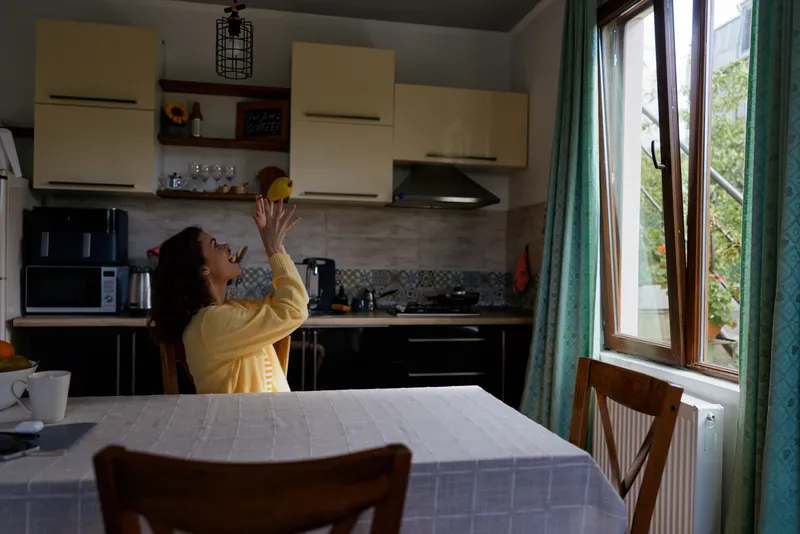 Cheerful woman in a bright kitchen reaching for a cabinet, candid lifestyle moment, warm natural light, cozy home atmosphere, everyday task, soft colors, relaxed posture, window view