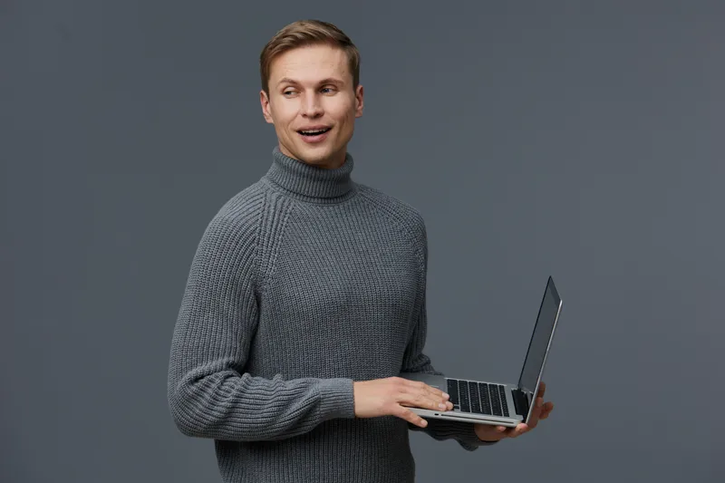 Smiling man holding laptop computer in hands, looking sideways, wearing cozy gray turtleneck, studio portrait on plain gray background, technology, work, casual style, indoor. People concept.