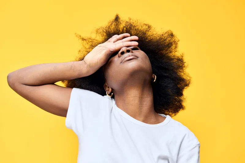 African American woman with curly hair expressing frustration against a bright yellow background, wearing a casual white t shirt, showcasing emotions of stress and overwhelm