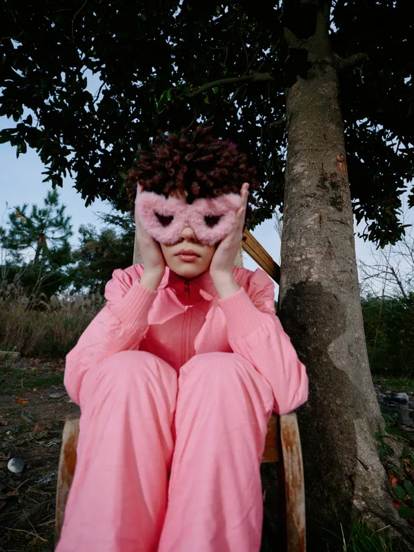 Pink attire, a lone person sits on a wooden chair beside a tree, holding a pink mask over the face, creating a mysterious, contemplative outdoor portrait with soft light