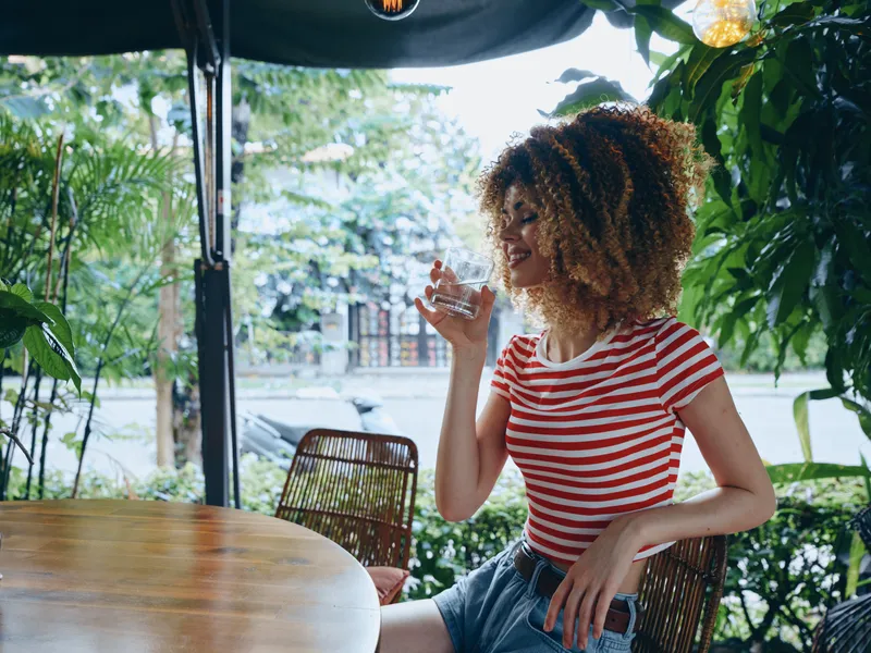 A young woman with curly hair and a red white striped shirt sits at an outdoor cafe table, smiling, holding a glass as sunlight and greenery frame the scene.