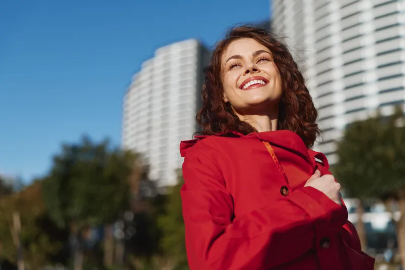 Smiling woman wearing a red coat stands outdoors in an urban setting on a sunny day, creating a cheerful, confident portrait with city high rises in the background