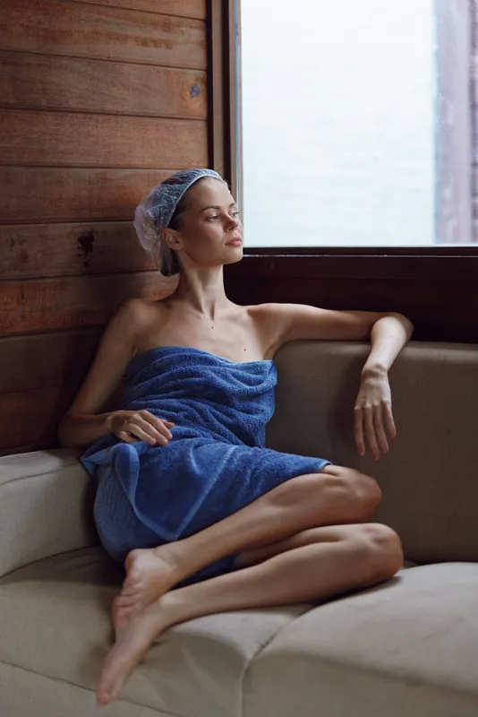Relaxed woman wrapped in blue towel sits on beige sofa, wearing shower cap near wooden wall and large window, enjoying peaceful moment of calm and self-care at home.