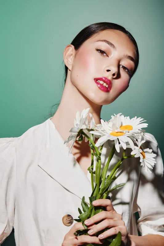 Young woman with red glossy lips holds white daisies wearing light jacket. Studio portrait on green wall. Concept for beauty, fashion, and floral creative photos.