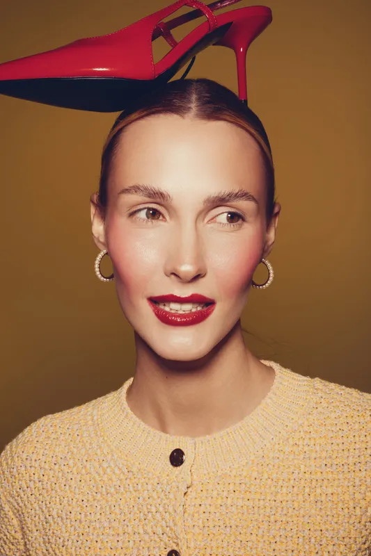 Young woman with red lipstick and hoop earrings balances a red high heel shoe on her head. She wears a yellow knitted sweater and looks to the side against a brown background.
