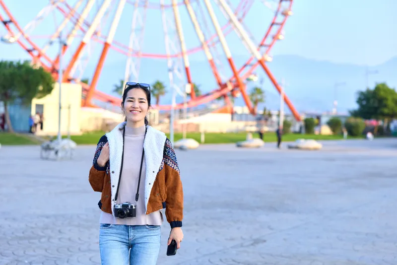 Smiling woman with a camera stands in a sunny amusement park with a large ferris wheel behind, casual fashion, open space, and a candid travel vibe conveying leisure and exploration