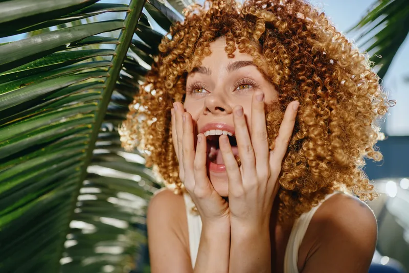 Excited woman with curly hair expressing surprise outdoors near tropical palm leaves under bright daylight with natural green background and warm tones.