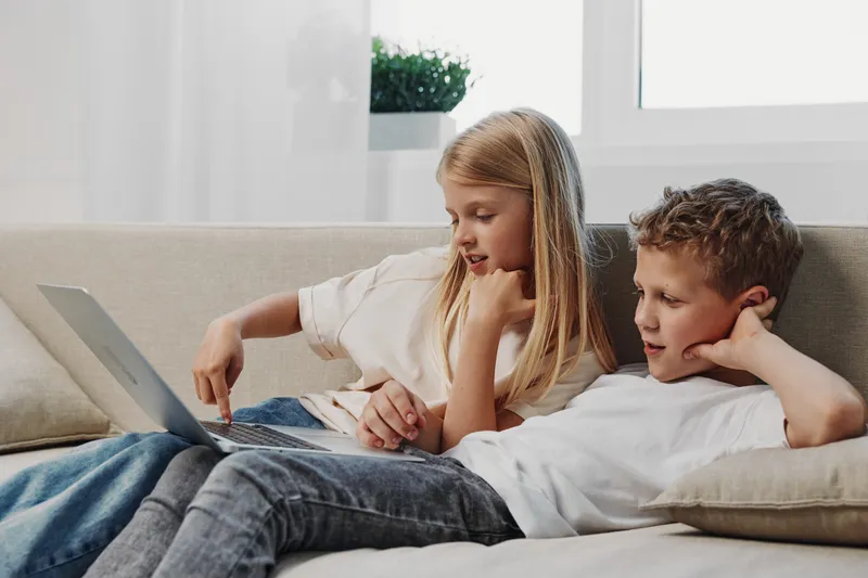 Two children sitting on a couch using a laptop together in a bright living room. The girl points at the screen while the boy watches attentively. Concept of learning and technology.