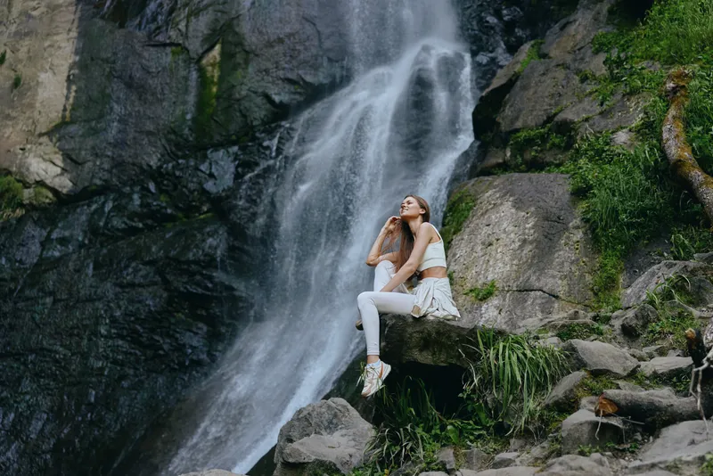 Young woman sitting near waterfall wearing white outfit, enjoying peaceful nature, surrounded by rocks and greenery, outdoor relaxation and calm atmosphere.