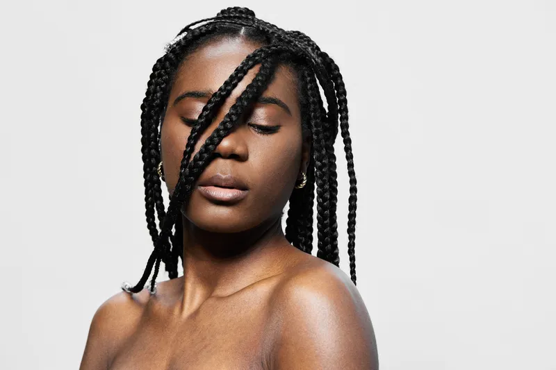 A young Black woman with braided hair, displaying a serene expression against a minimalist white background, exuding confidence and elegance