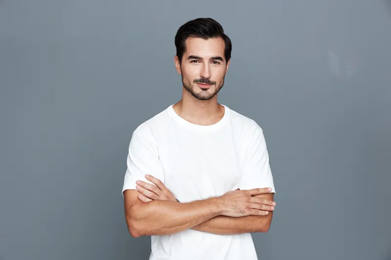 Confident smiling man wearing casual white t-shirt stands with arms crossed against a gray background showcasing a modern and stylish portrait. Men lifestyle concept.