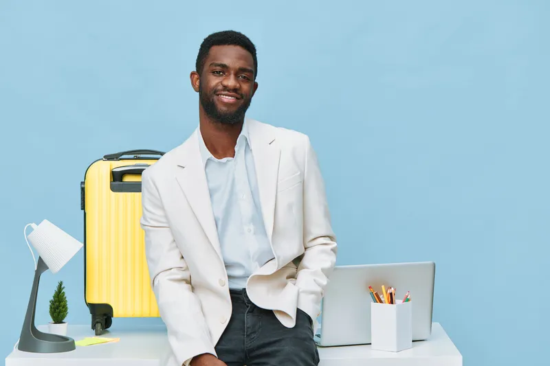 Smiling man in light suit leaning on desk with laptop and suitcase against blue background, professional travel or business concept, casual style, positive mood, modern setting.