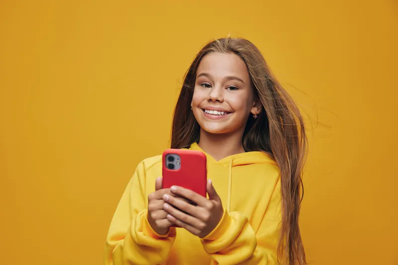 Girl in yellow hoodie smiling and holding red smartphone for communication and social networking on orange background. Youth, technology and casual style with happy expression.