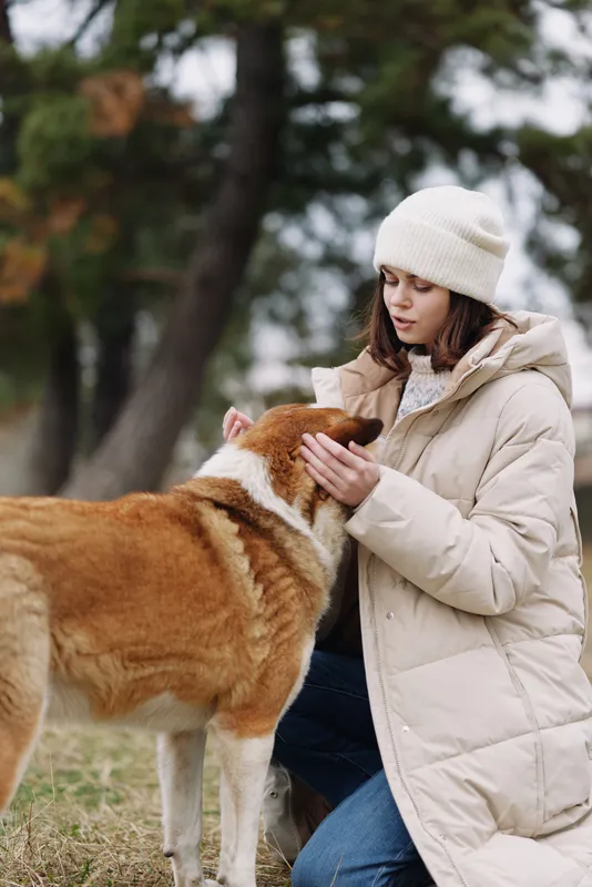 A woman in a winter coat and beanie gently pets a dog outdoors in a park, capturing a calm, affectionate moment of companionship and bond between person and pet in a natural setting.