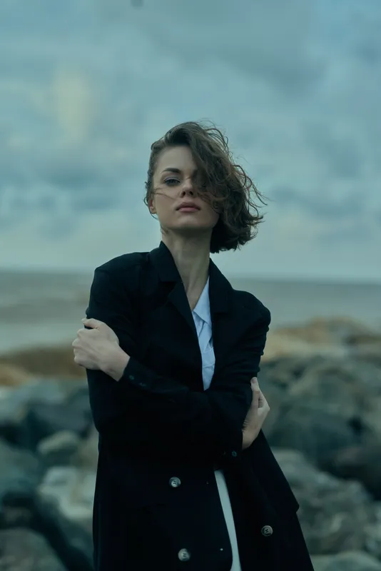 Woman on a windy rocky shoreline wearing a dark coat and light shirt, arms crossed, windswept hair, moody sea backdrop, fashion and outdoor style for editorial or travel vibe in dramatic composition