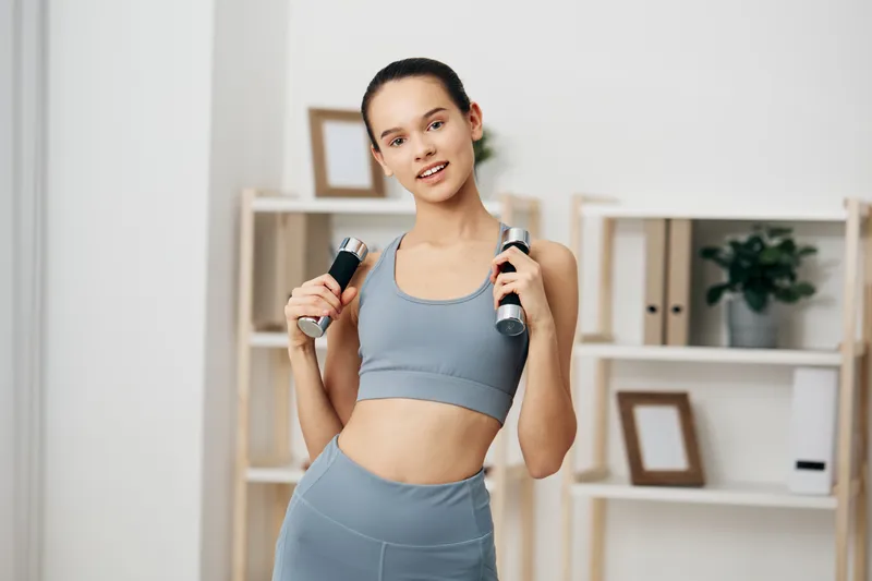 Woman fitness sport exercise with dumbbells in a bright home gym, wearing teal athletic wear, mid-workout, showing focus and energy during a strength training session.