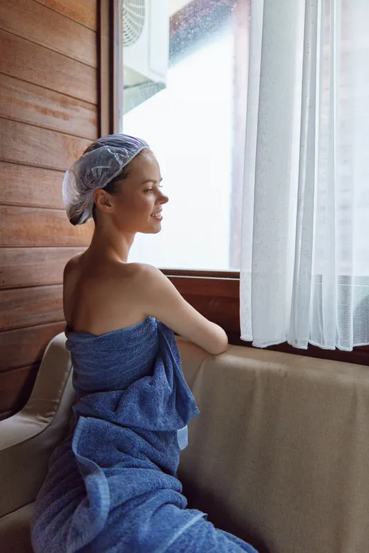 woman in spa with towel and shower cap sitting near window, enjoying natural light and peaceful wooden interior atmosphere, wellness and relaxation concept