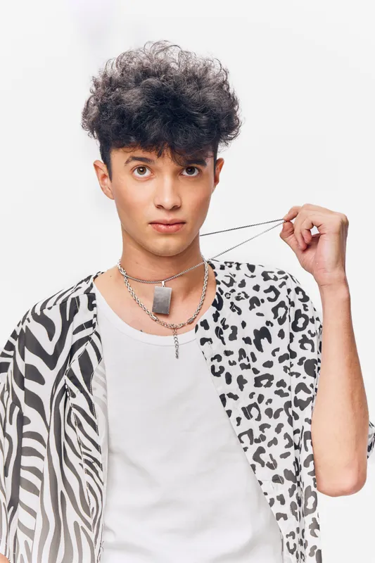 Young man with curly hair wearing a patterned shirt and holding a chain necklace, posing confidently against a plain white background.