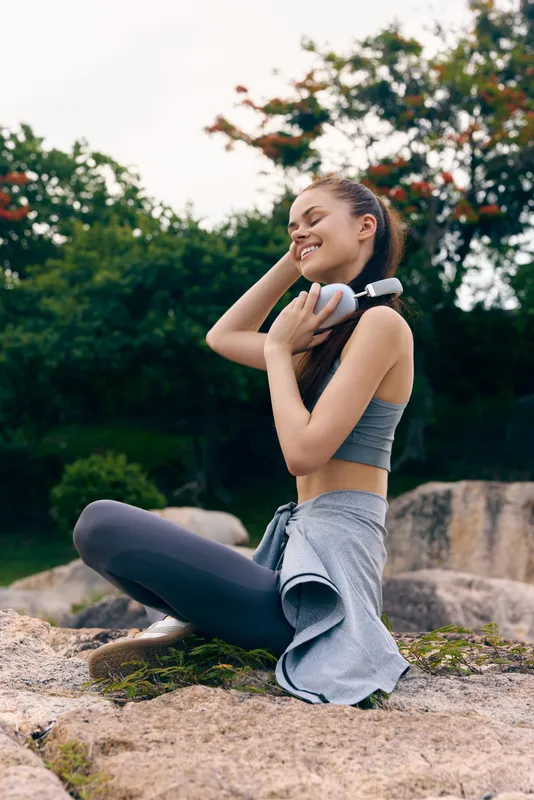 Young woman enjoying music outdoors in activewear, smiling joyfully while sitting on rocks in a natural green setting