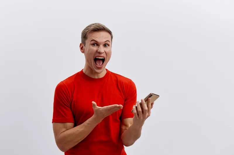 Excited smiling man in red t-shirt reacting with surprise while looking at smartphone, gesture of amazement and joy, indoors with plain white background, casual style, human emotion concept