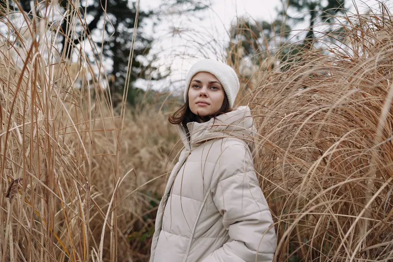 Winter scene with a woman in a beige coat standing among tall grasses in an open field, calm pose, soft light, cozy fashion portrait for lifestyle and outdoor wear