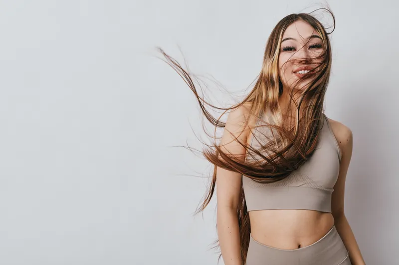 Young woman with long hair smiling and wearing beige sportswear on gray background, motion captured with hair flying, healthy lifestyle and fitness concept.
