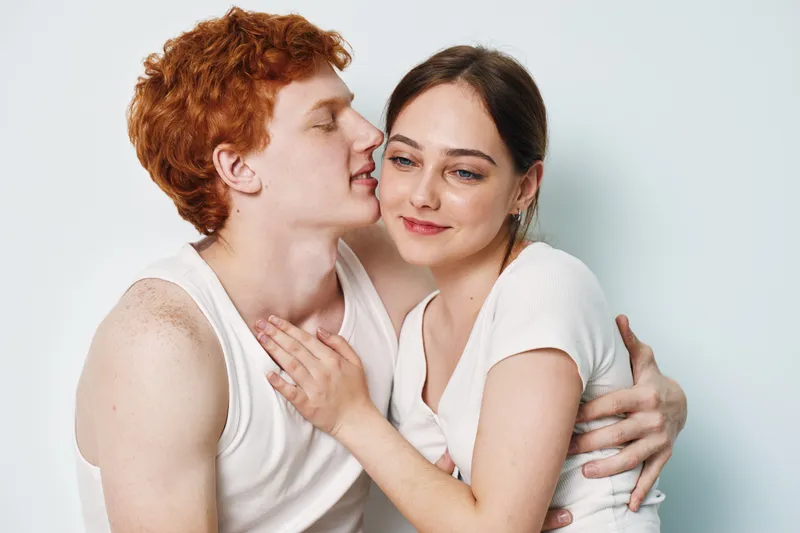 Couple, love and affection with young man whispering to woman in tender embrace for romance, intimacy and relationship on white background at studio. Emotional connection between two people.