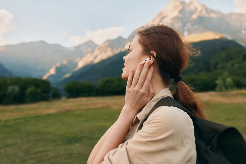 Young woman with headphones stands in a grassy field, backpack on her shoulder, mountains in the distance as she savors a calm outdoor moment