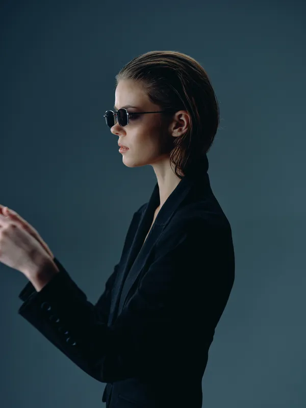 Stylish woman in black suit wearing sunglasses with wet hair against dark background, looking thoughtful and confident.