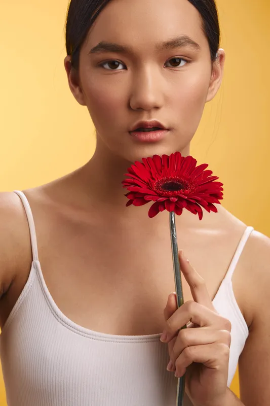 Asian woman poses in a studio portrait, holding a red flower with a gentle smile and calm gaze, creating a warm, elegant mood in a minimal setting.