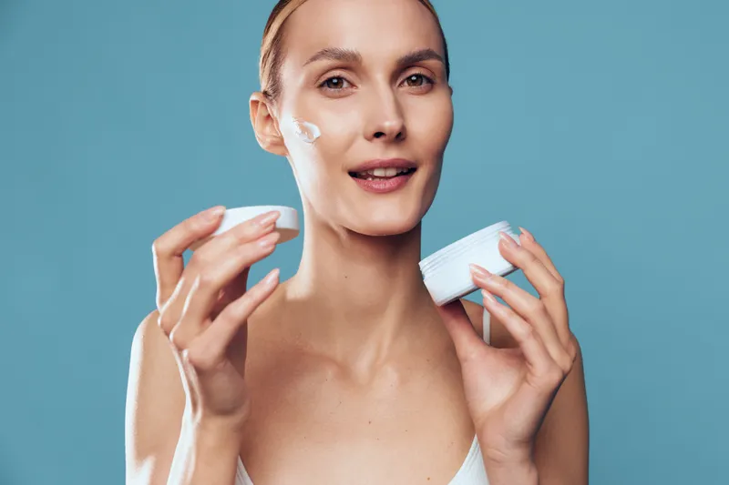 A young woman applies moisturizer to her face with a gentle smile in a bright studio, conveying a calm skincare moment and self care routine.