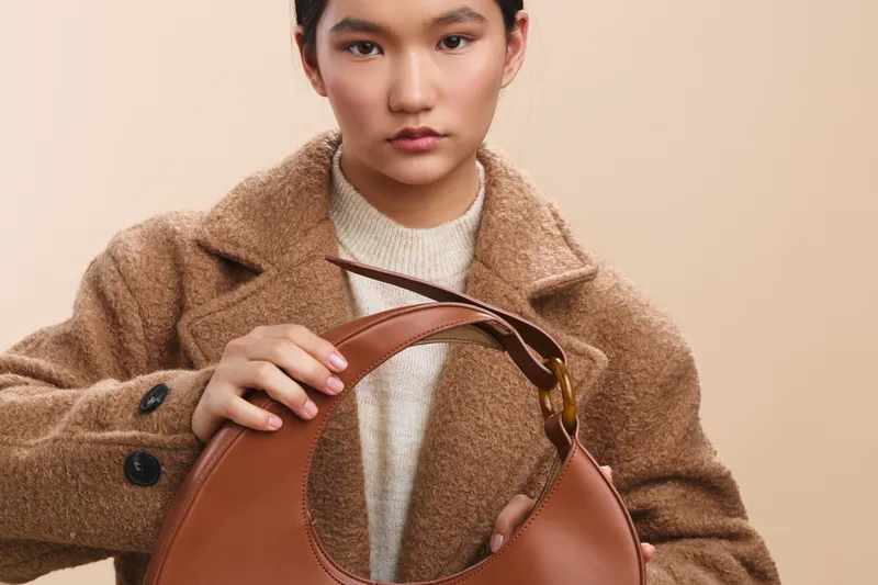 asian woman wearing a cozy coat examines a brown handbag against a warm beige background, creating a stylish fashion moment with a calm, focused expression and subtle texture.
