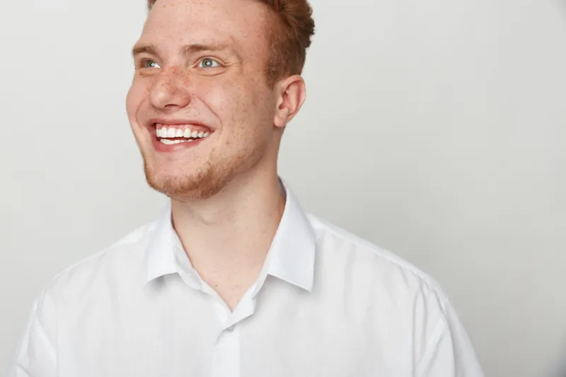 Smiling young man with red hair and beard wearing white shirt looks away happily on light background. Concept of positive emotions, joy, and casual lifestyle portrait.