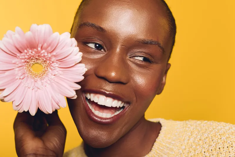 Smiling dark skinned African woman holds pink flower near face on yellow background. Concept of natural beauty, happiness, skincare, and floral wellness for black female model.