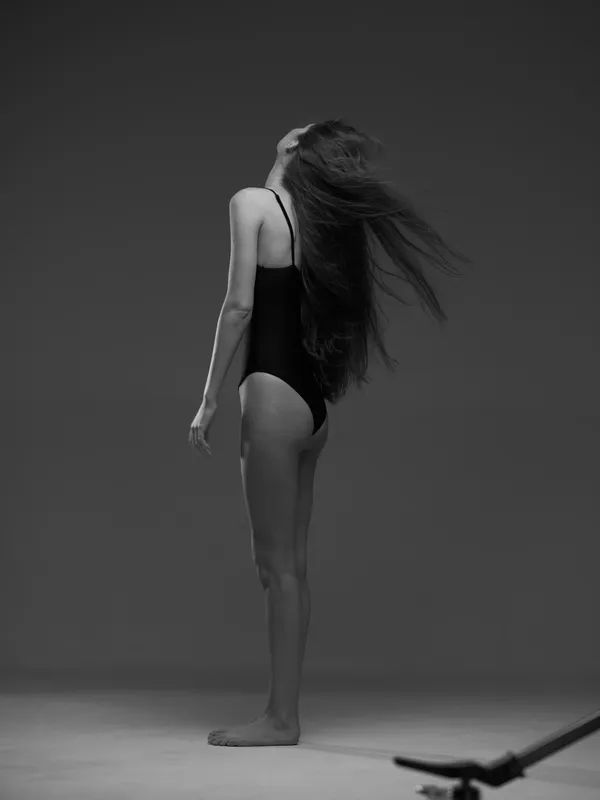 Black and white woman standing with long hair flowing wearing a black swimsuit against gray background