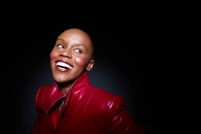 Smiling African American woman with dark skin wearing a shiny red leather jacket looks away from camera on black background. Concept of joy, fashion, confidence, and beauty portrait.