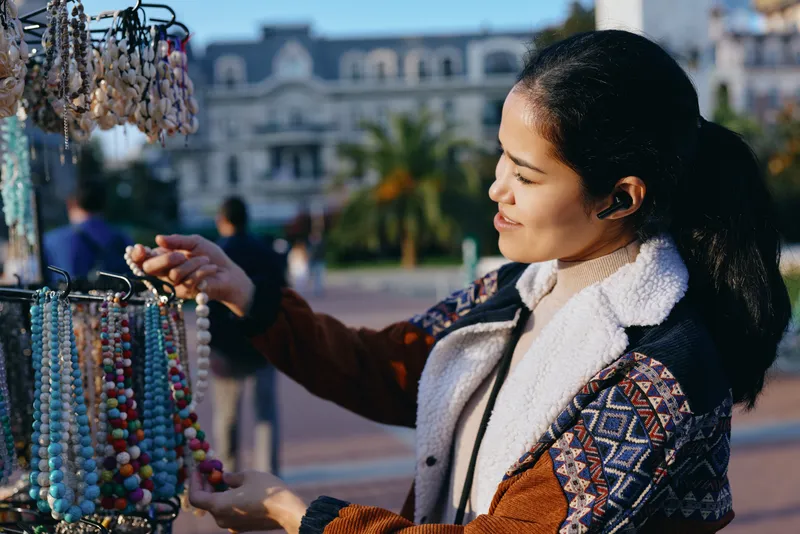 Woman browses colorful beads at outdoor market, selecting jewelry accessories in a sunny plaza, fashion display with vibrant string beads and handmade ornaments
