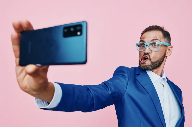 Professional businessman in a blue suit takes a selfie against a pink background, exuding confidence and surprise. Isolated colorful backdrop highlights a modern, dynamic corporate vibe.