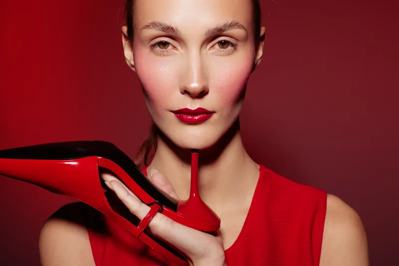 Close-up of a woman with red lipstick and blush holding a red high heel shoe near her face against a red background. Concept of fashion, beauty, style, and elegance.