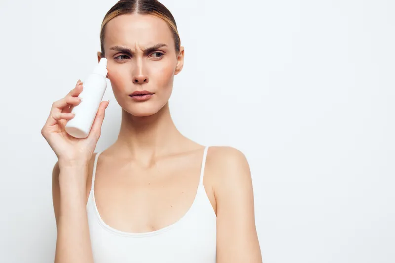 Young woman with blonde hair in a white tank top applies facial mist on her face with a focused expression against a white background for skincare and beauty concept.