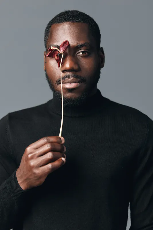 African man with dark skin holding dried flower in front of eye in creative conceptual visual style. Portrait of black male model wearing black sweater with artistic expression on gray background.