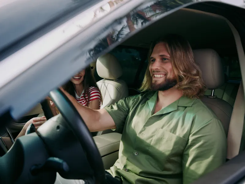 A smiling man with long hair drives a car in a green shirt, while a cheerful passenger sits beside him; a candid moment of road trip joy and companionship.
