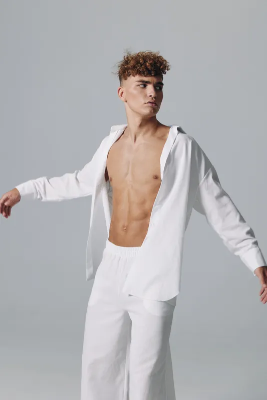 Young man with curly hair wearing white pants and open shirt posing with arms outstretched on gray background in studio. Fashion, style and casual clothing concept.